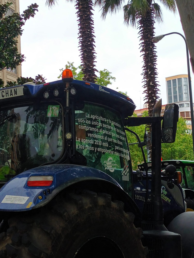Un tractor en plena vía urbana muestra un mensaje en la ventanilla: "La agricultura y la ganadería son los únicos sectores en los cuales compramos al precio que nos dicen y vendemos al que nos dicen. La agenda 2030 esta... para enriquecer a los ricos y empobrecer a los más pobres".