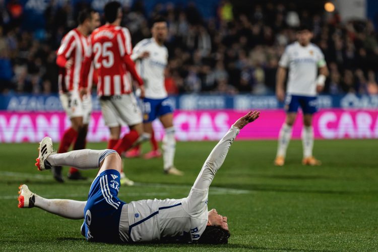 Un jugador del zaragoza se retuerce de dolor en el suelo y detrás aparecen varios jugadores del Sporting conversando con los del Zaragoza.